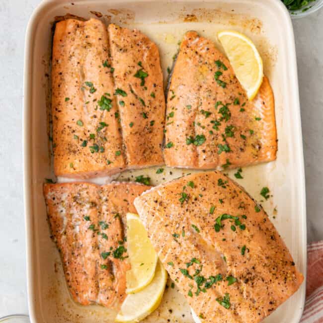 Fish spatula lifting up one lemon pepper salmon from baking dish with 3 more fillets garnished with lemon wedges and fresh parsley.