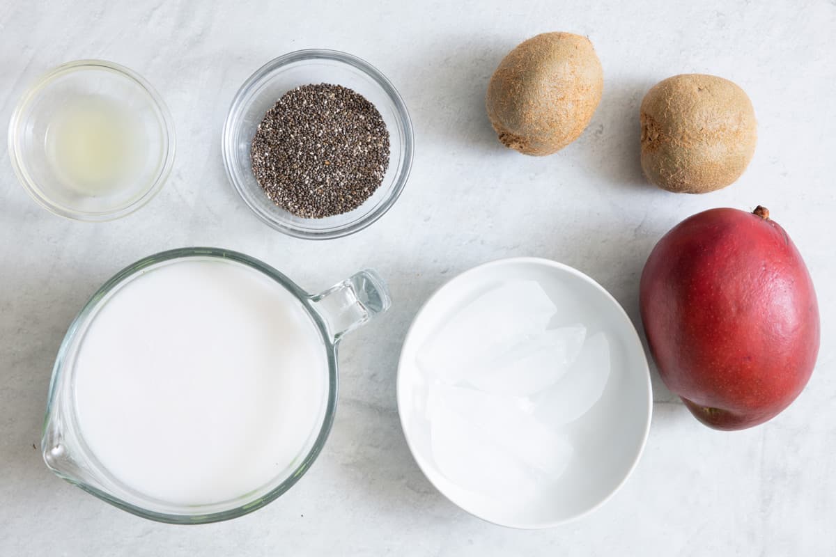 Ingredients for smoothie before being prepped: lemon juice, chia seeds, 2 whole kiwis, 1 whole mango, bowl of ice, and cup of milk.