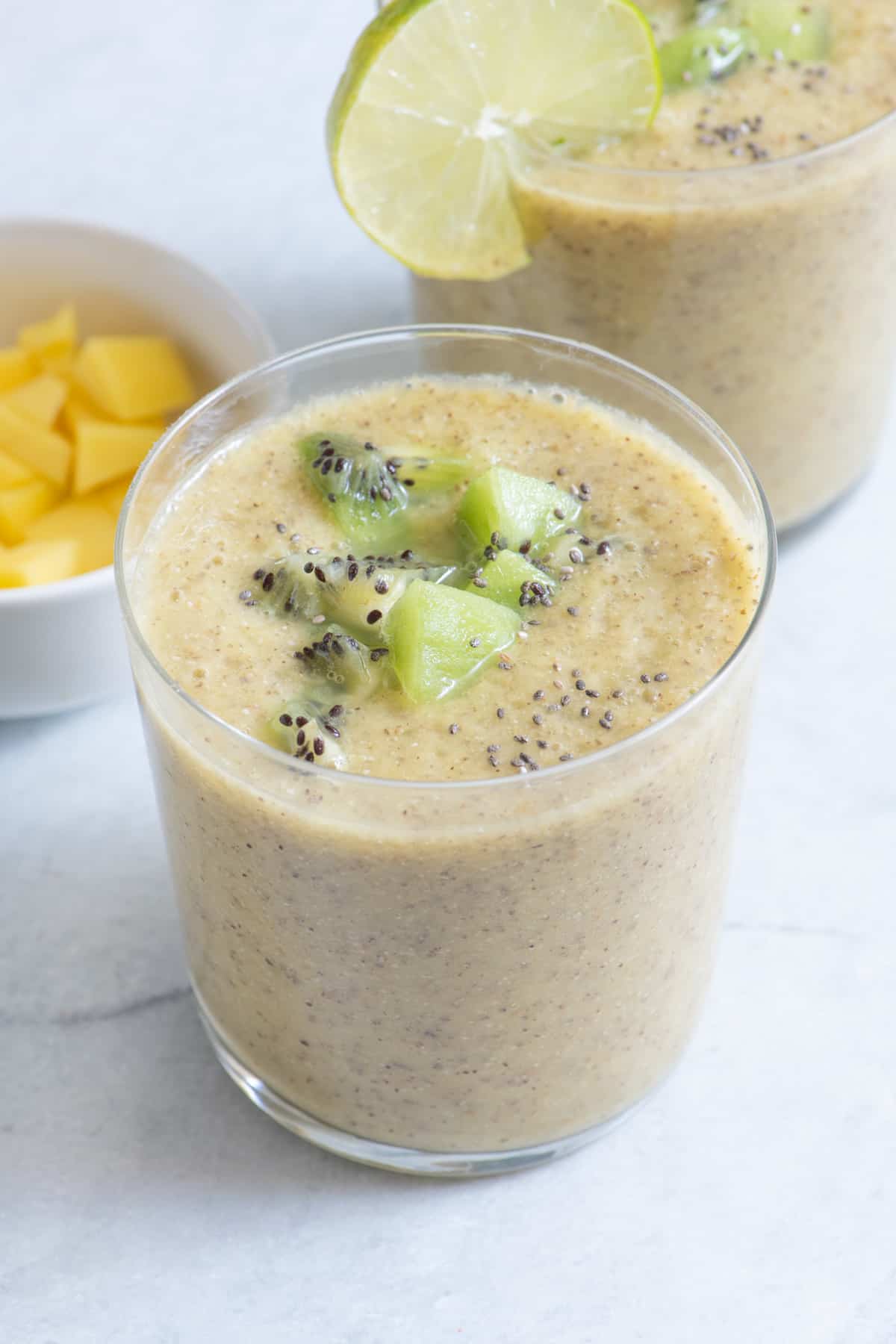 2 small glasses of mango kiwi smoothie with extra chunks of mango and kiwi on top and a round lemon slice on 2nd glass.