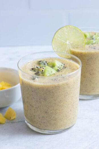 Two glasses of mango kiwi smoothie with chunks of fresh kiwi on top, a bowl of mangos on the side, and a lemon slice on second glass.