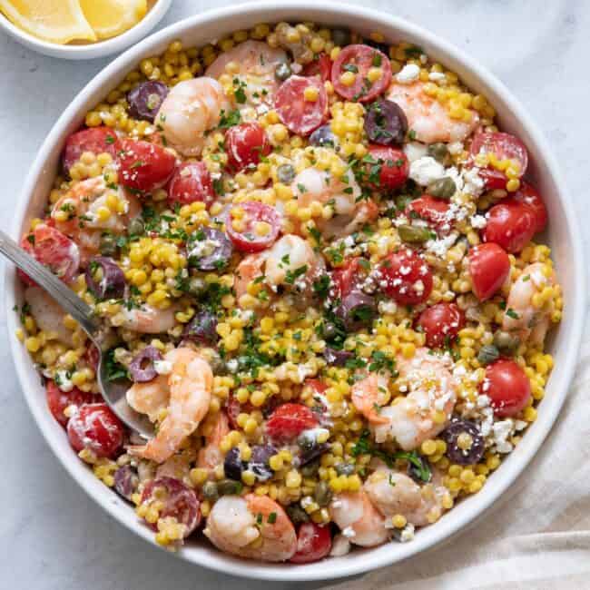 Close up shot of the Mediterranean Couscous in large white serving bowl with chopped parsley and lemon wedges off to side.