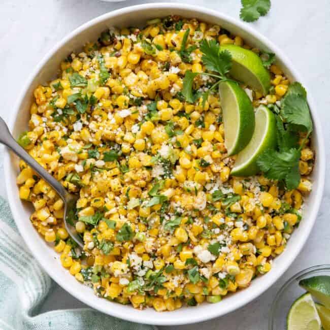 Overhead shot of Mexican Street Corn Salad in large white bowl with lime wedges and side of cotija cheese crumbles.