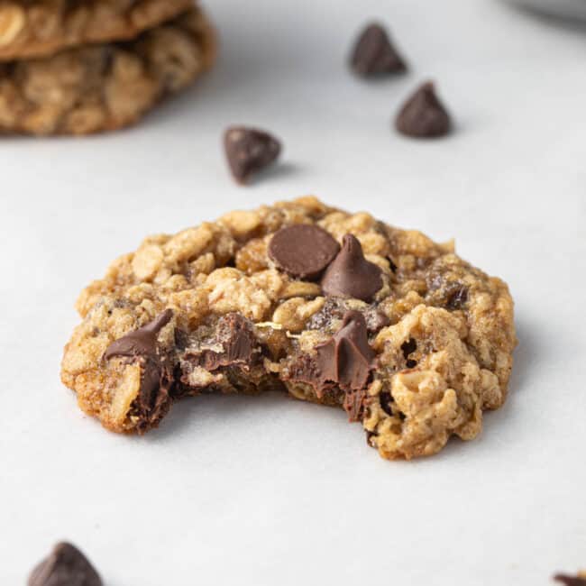 Stack of oatmeal cookies with chocolate chips on parchment paper with one cookie infront with bite taken out and a small dish of chocolate chips in the back.