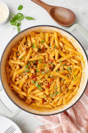 One Pot Turkey Pasta in pot, garnished with parmesan cheese and fresh basil with a wooden spoon nearby.