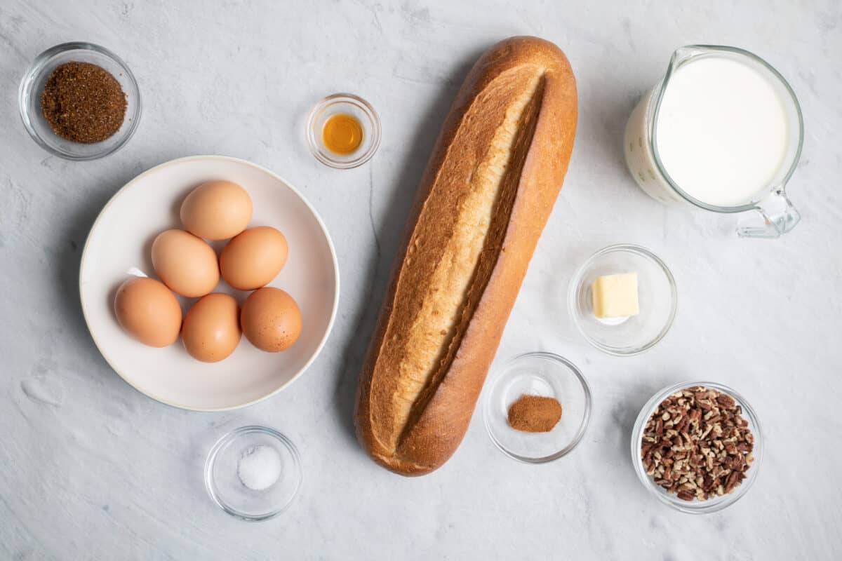 Ingredients for recipe before prepped in individual bowls and cups: dark brown sugar, 6 whole eggs, salt, vanilla, loaf of french bread, cinnamon, butter, milk, and chopped pecans.