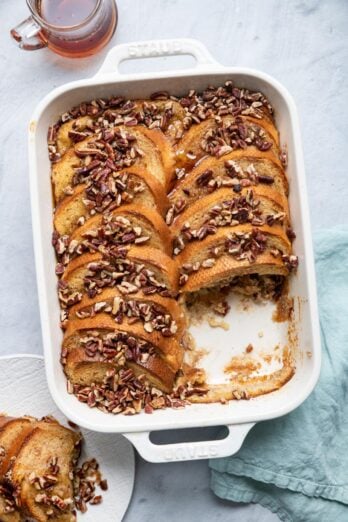 Baked french toast topped with chopped pecans in a large white baking dish and a portion removed on a plate nearby with maple syrup on top.