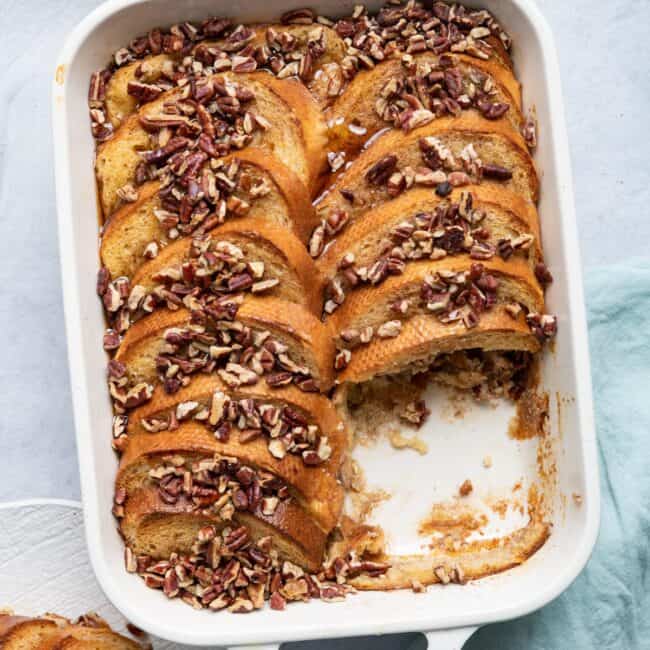 Baked french toast topped with chopped pecans in a large white baking dish and a portion removed on a plate nearby with maple syrup on top.