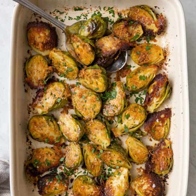 Parmesan crusted brussel sprouts in a baking dish flipped cut side up with crusted cheese with a serving spoon tucked and garnished with fresh parsley and flaky salt.