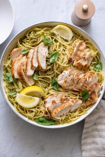 Finished pasta in dish tossed in pesto and topped with sliced chicken, basil, and lemon.