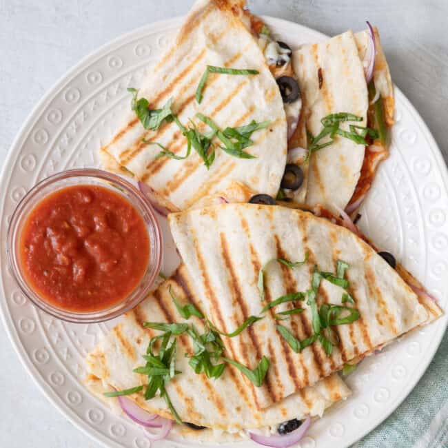 Recipe cut into hand held sizes on a round white plate with a side of marinara sauce for dipping.