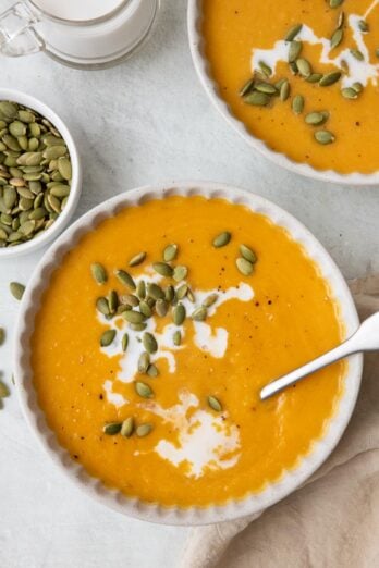 Bowl of soup garnished with coconut milk and pumpkin seeds with spoon inside bowl and another bowl nearby.