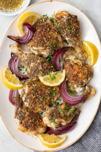 Roasted Zaatar chicken arranged on an oval platter with seasoned skin, garnished with red onion slices, lemon wedges, and fresh parsley.