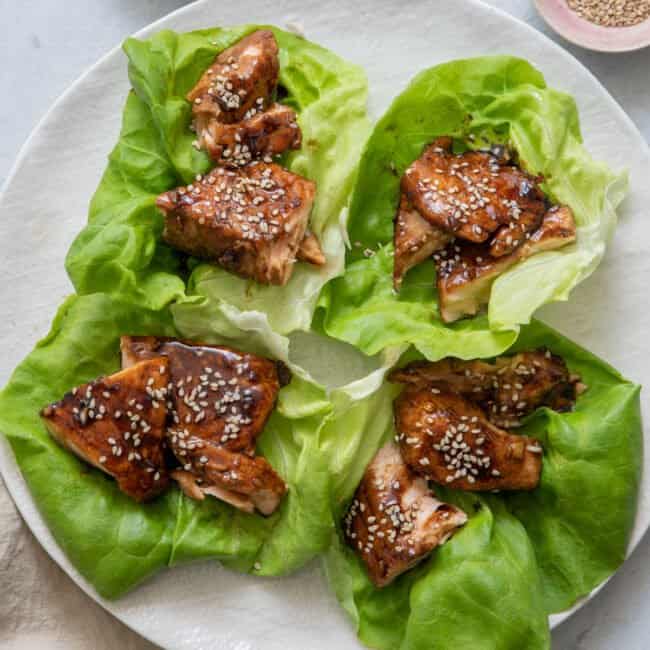 Four Salmon Lettuce Wraps on large white plate with a side dish of both scallions and sesame seeds.