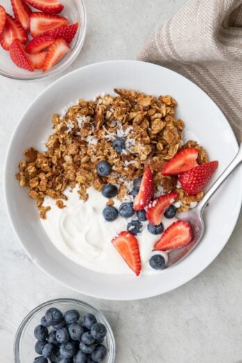 Bowl with scrambled oats, greek yogurt, a few quartered strawberries, blueberries, with coconut sprinkled on top. Small dishes of berries nearby.