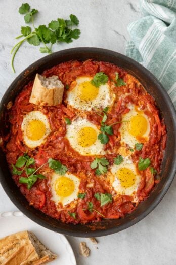 Shakshuka in pan garnished with cilantro and sourdough nearby with a piece in on top and showing cooked eggs in rich sauce.