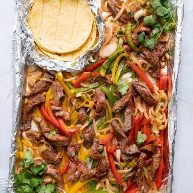 Sheet pan with steak fajita ingredients tossed together, garnished with fresh cilantro with a side of corn tortilla shells in an open foil pack.