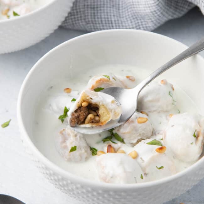 Spoon lifting up a dumpling from a bowl of Shish Barak that is cut open to show beef and nut mixture inside.