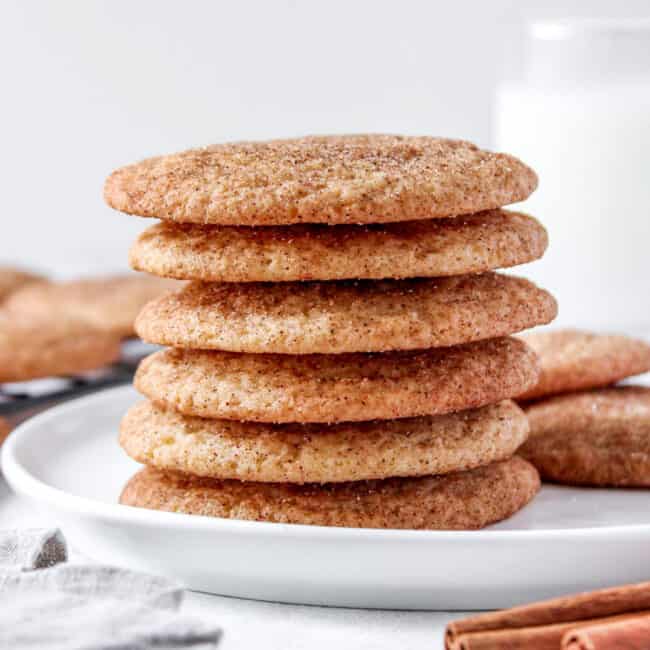 Snickerdoodles stack tall on a round plate with extra cookies nearby and a glass of milk in the background.