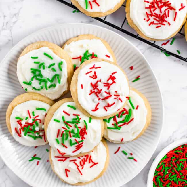 Soft baked Lofthouse cookies topped with icing and sprinkles on a round plate next to a cooling wire rack with more cookies. Small bowls of icing and sprinkles nearby.