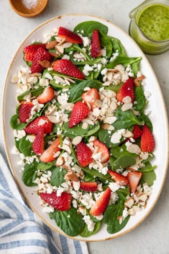 Spinach strawberry salad on a large oval platter topped with feta and sliced almonds with a basil vinaigrette nearby.