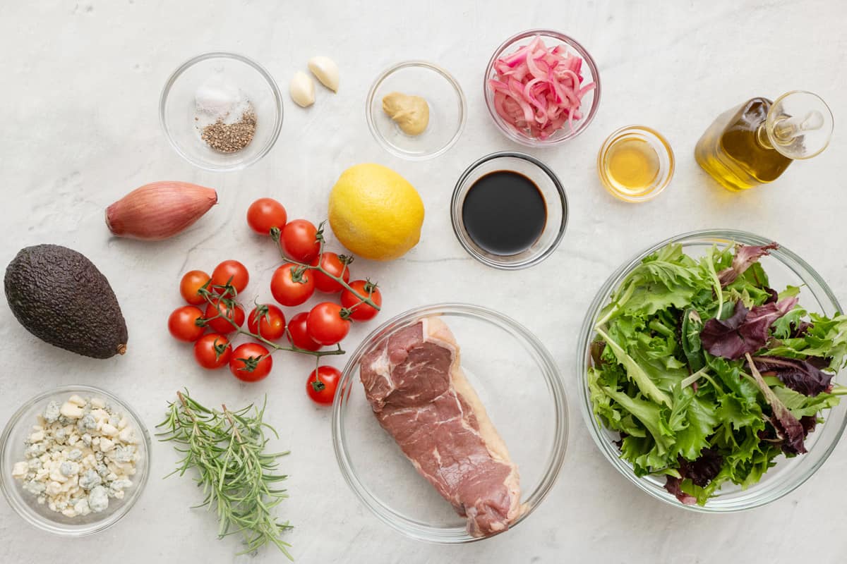 Ingredients for recipe in individual bowls and before prepped: crumbled blue cheese, avocado, shallot, seasoning, garlic cloves, vine tomatoes, lemon, Dijon, balsamic vinegar, honey, oil, and spring mix.