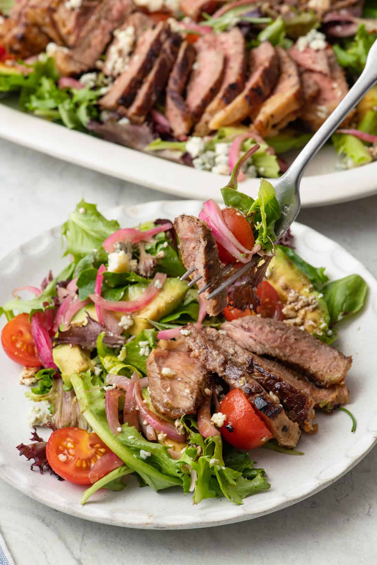 Fork lifting up a bite of steak salad from a small serving plate with platter of recipe in background.