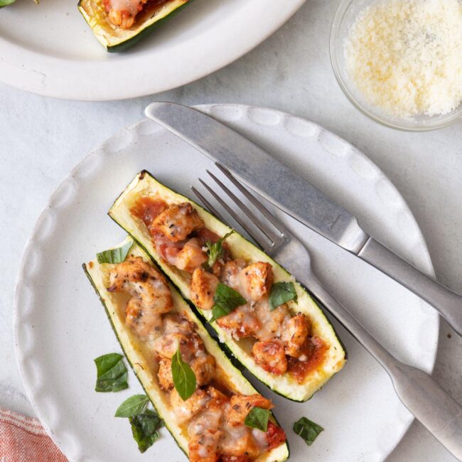 Final plating of the stuffed zucchini boats topped with fresh basil with fork and knife on plat and a side of grated parmesan to the side.