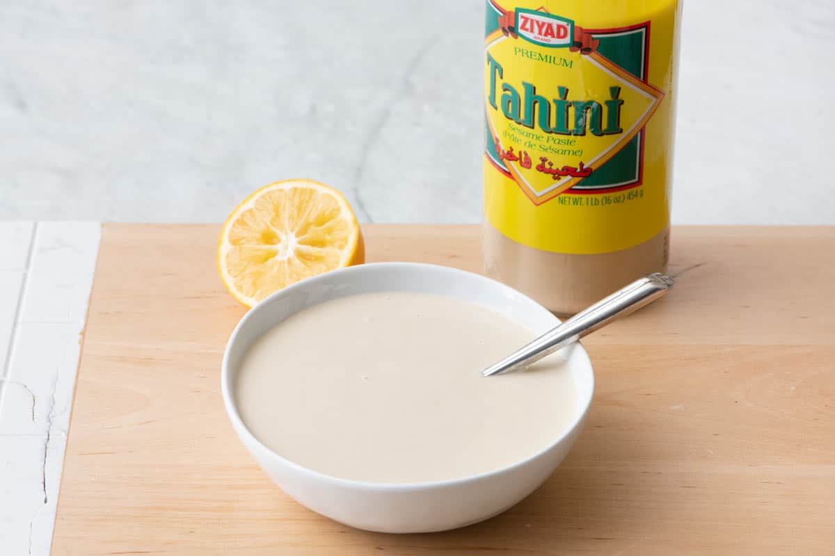 Bowl of tahini sauce next to a jar of Ziyad tahini sesame seed paste and a halved lemon.