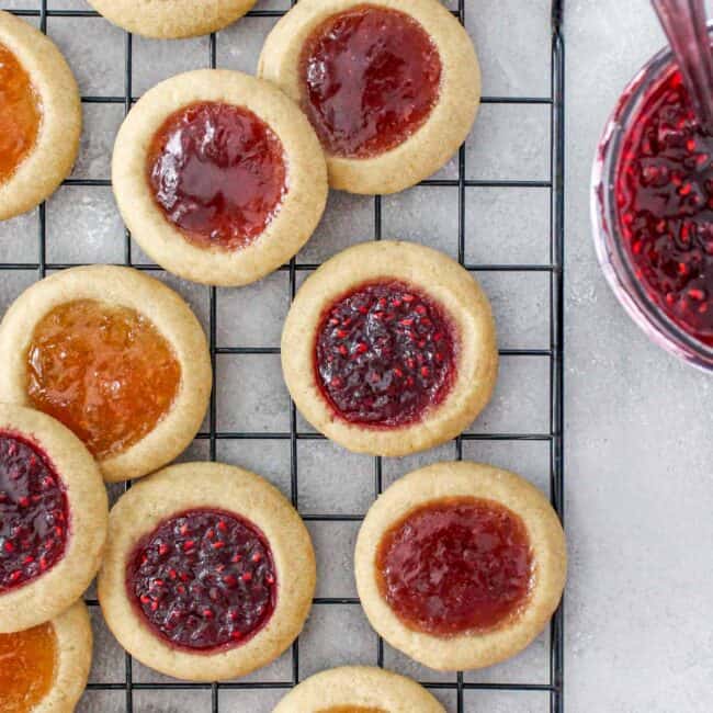 Three different jam filled thumbprint cookies on a wire rack with a jar of jam nearby.