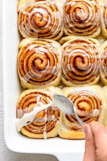 Spoon glazing vegan cinnamon rolls in white pan