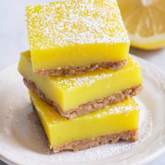 Stack of three vegan lemon bars on small white dish with half lemon in background