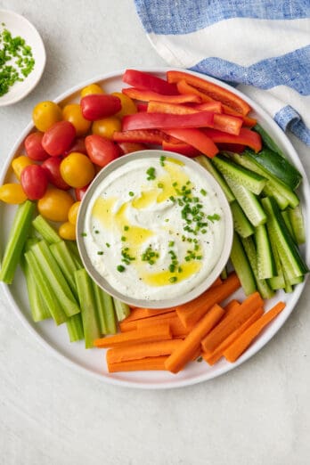 Whipped cottage cheese in a bowl garnished with chives and olive oil. Bowl on a platter surrounded by fresh veggies cut into sticks, including red bell peppers, zucchini, carrots, celery, and tomatoes.